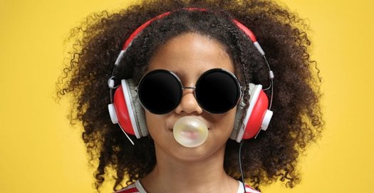 Ein Mädchen mit Sonnenbrille und rot-weißen Kopfhörern bildet mit ihrem Kaugummi eine Blase. Der Hintergrund ist gelb