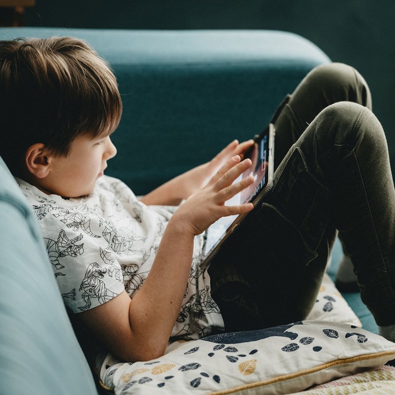 Ein Junge sitzt mit einem Tablet auf dem Schoß auf einem hellblauen Sofa